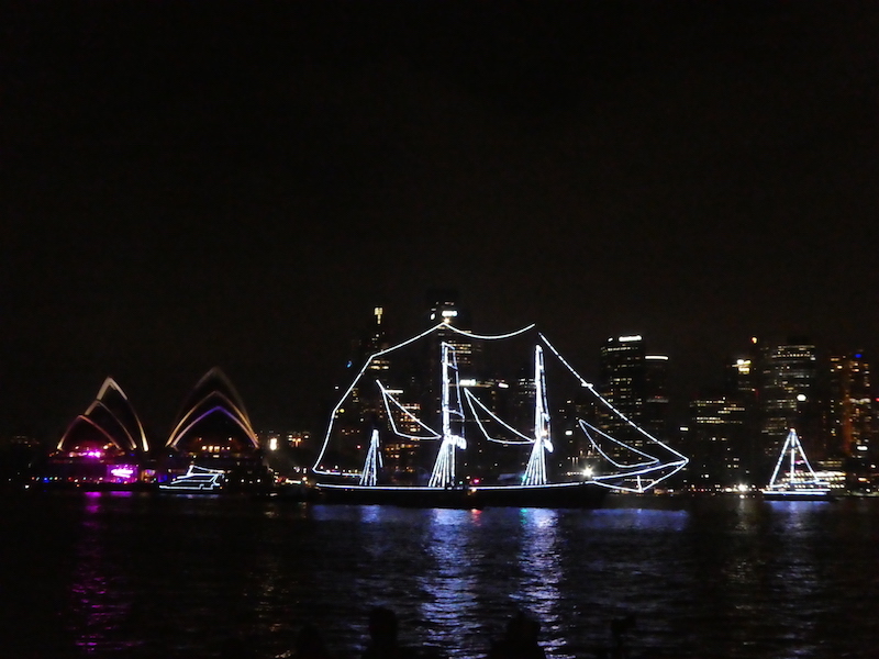Na pierwszym tle podświetlony żaglowiec w tle centrum biznesowe Sydney i Opera